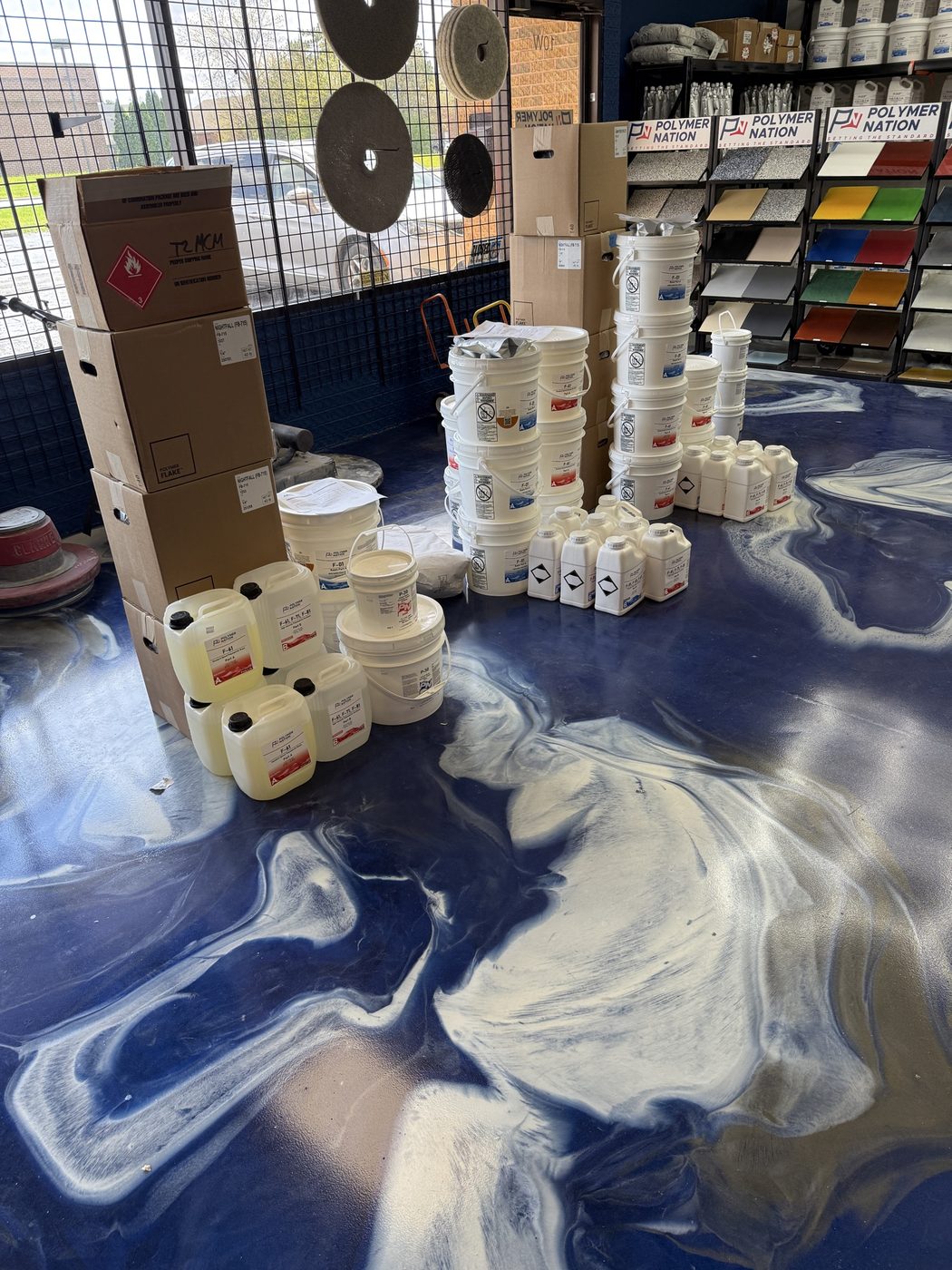 A staged pickup order at the ATC Supply store: stacked resin pails, jugs of hardener, flake boxes, buff pads, and a floor buffer lined up by the front bay window ready for a contractor to load out