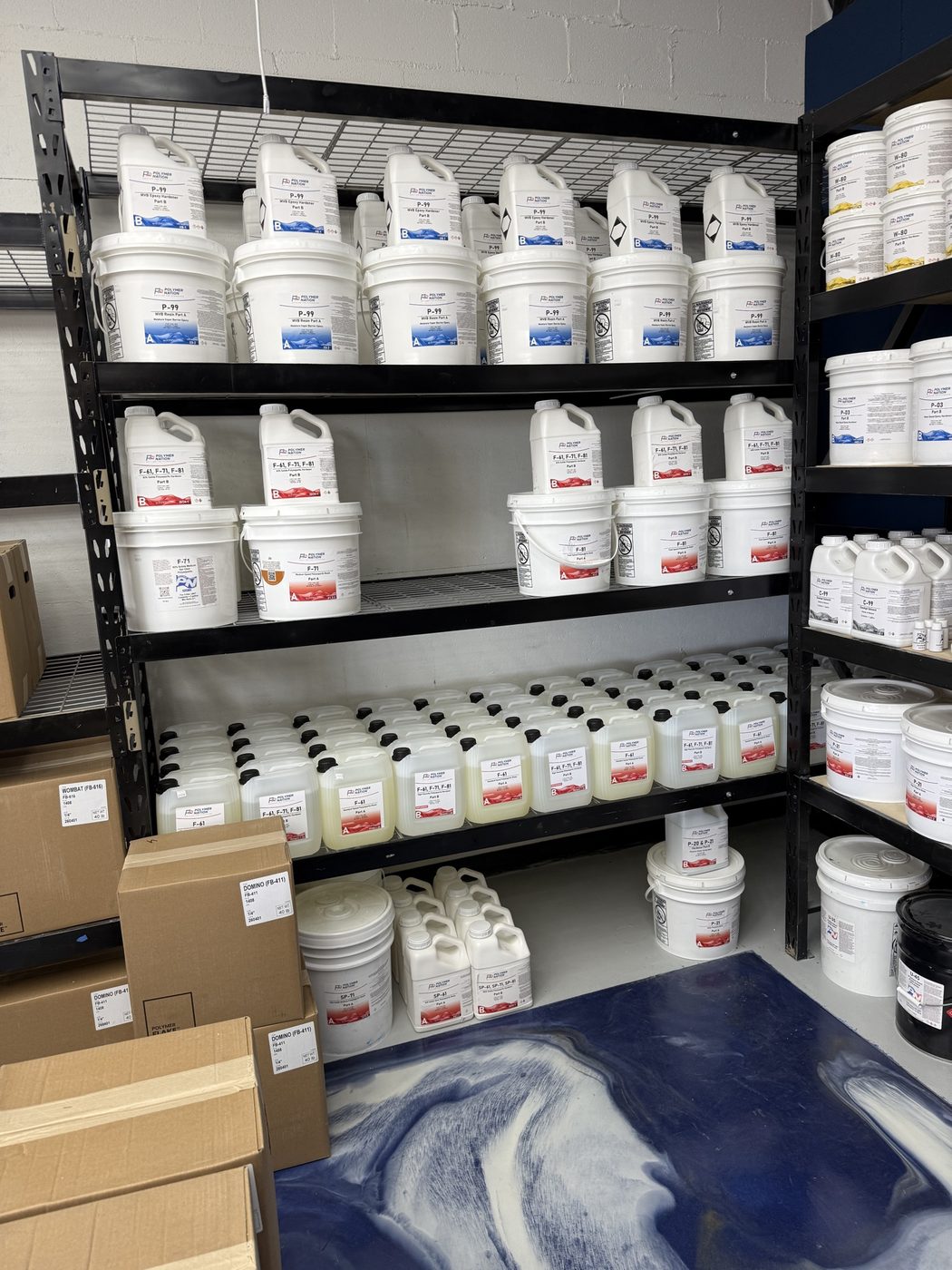 Deep stack of Polymer Nation P-99 epoxy resin and hardener pails alongside F-41, F-71, F-81 topcoat containers filling the shelving from floor to ceiling