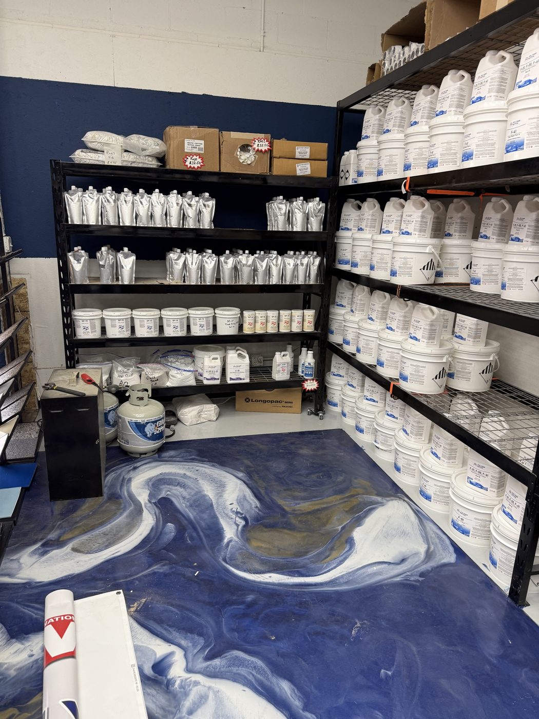 Polymer Nation polyaspartic pouch and urethane pail shelving corner with propane tank and broadcast tools on the metallic navy floor