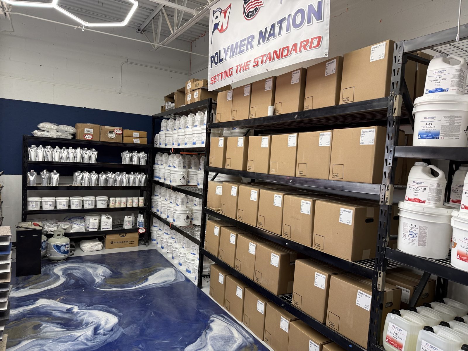Wide view of the ATC Supply stockroom showing the Polymer Nation flake box wall, silver polyaspartic pouch shelving, resin pails, and Polymer Nation Setting the Standard banner overhead