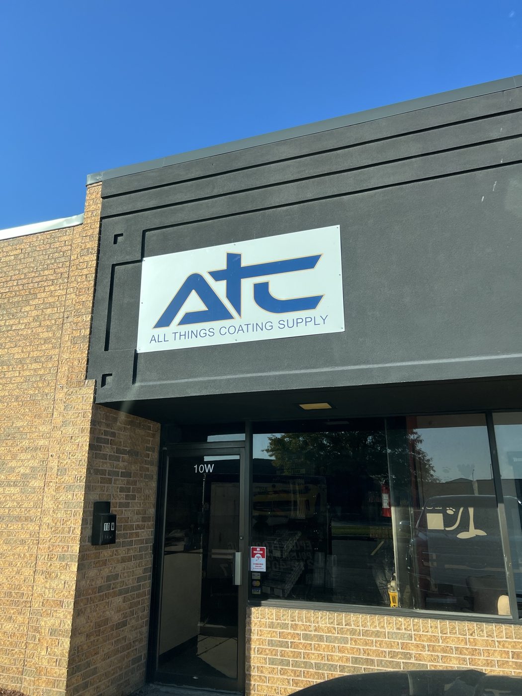 ATC — All Things Coating Supply exterior signage above the entrance to unit 10W in Merrillville, Indiana, shown in golden-hour light against brick and charcoal facade