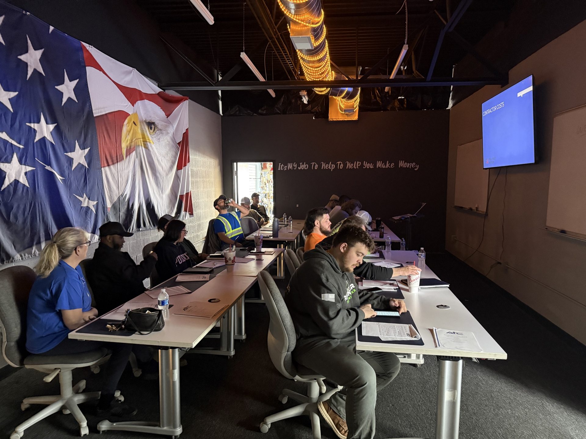 Contractors seated at ATC Supply training class studying contractor cost sheets in front of a large American flag mural